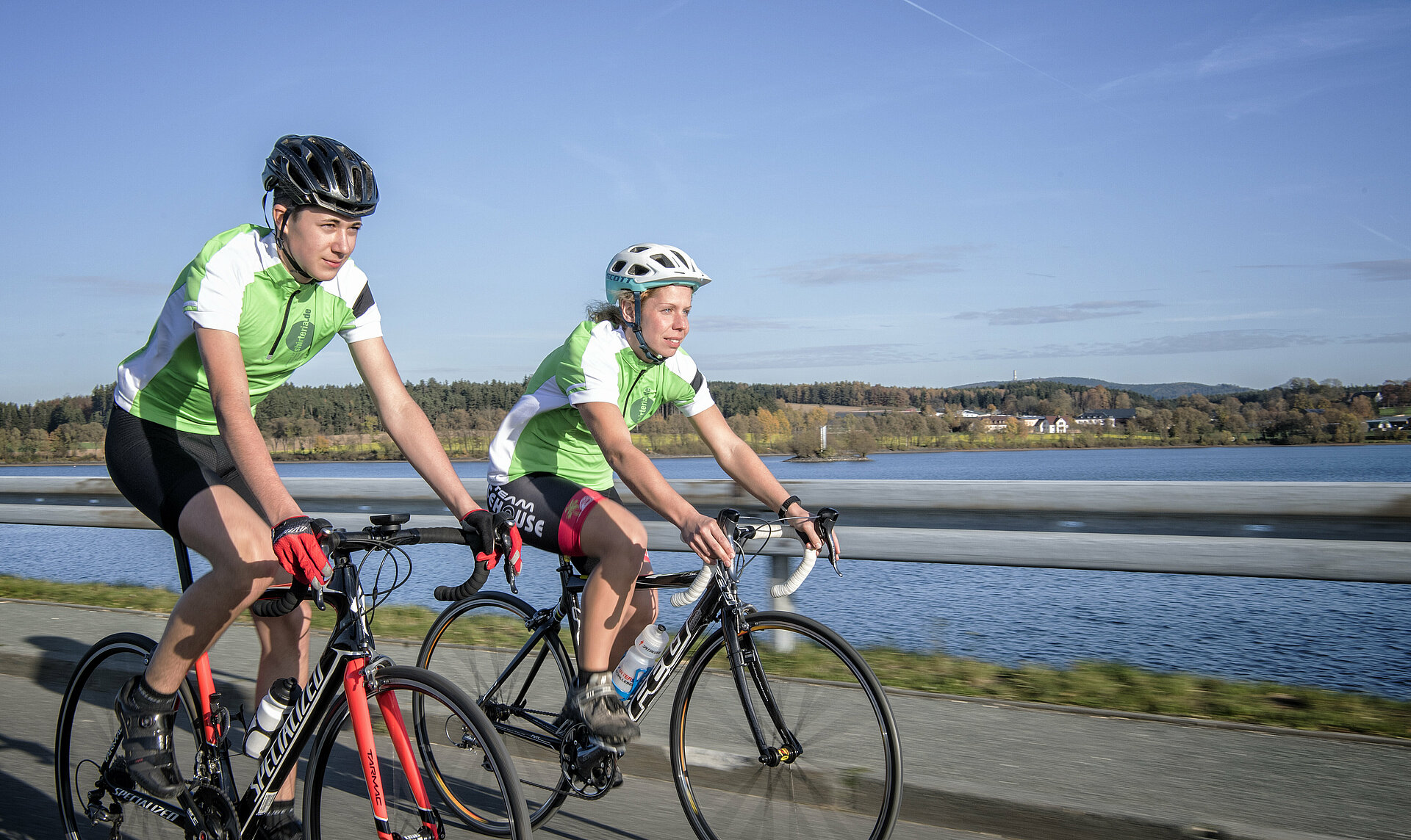 Zwei Rennradfahrer in voller Fahrt vor einem See und blauem Himmel