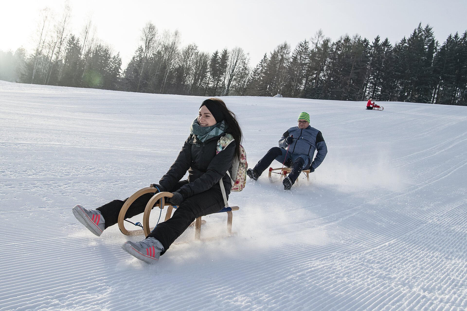 Blick auf den Rodelhang. Drei Personen fahren mit dem Schlitten ab. Dabei stiebt der Schnee auf.