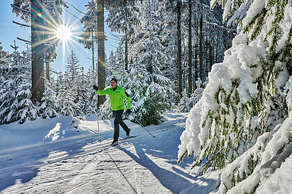 Langläufer im tief verschneiten Winterwald. Die Sonne strahlt durch die Bäume im Hintergrund