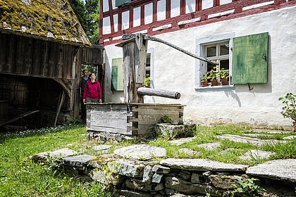 Zwei Personen vor einem Fachwerkgebäude mit Brunnen und Scheune