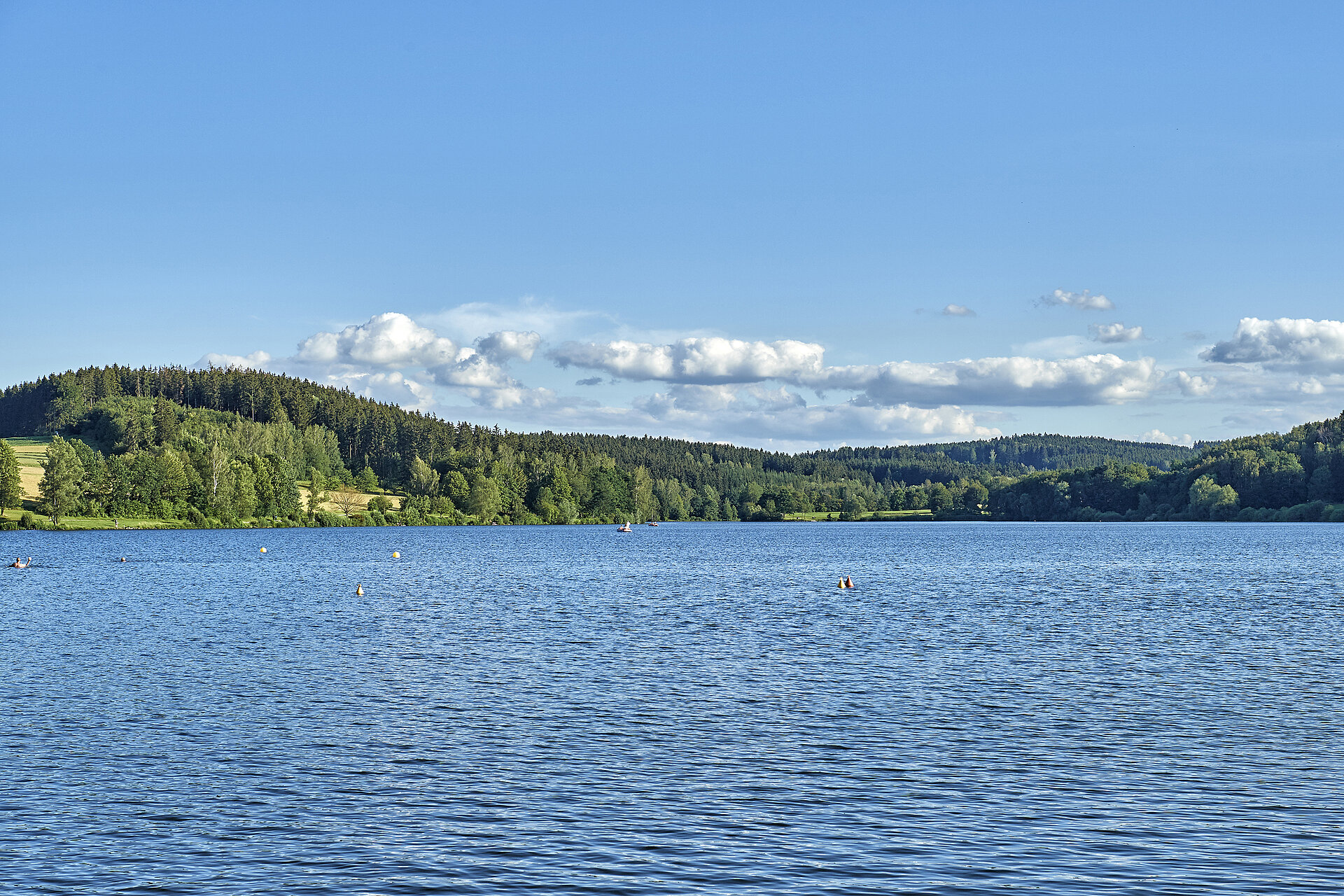 Im Vordergrund der See, Im Hintergrund grüne Hügel und blauer, leicht bewölkter Himmel