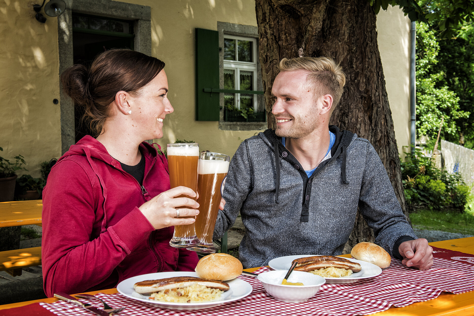 Ein Mann und eine Frau sitzen im Biergarten an einem gedeckten Tisch. Sie haben Biergläser in der Hand uns stoßen gerade an. 
