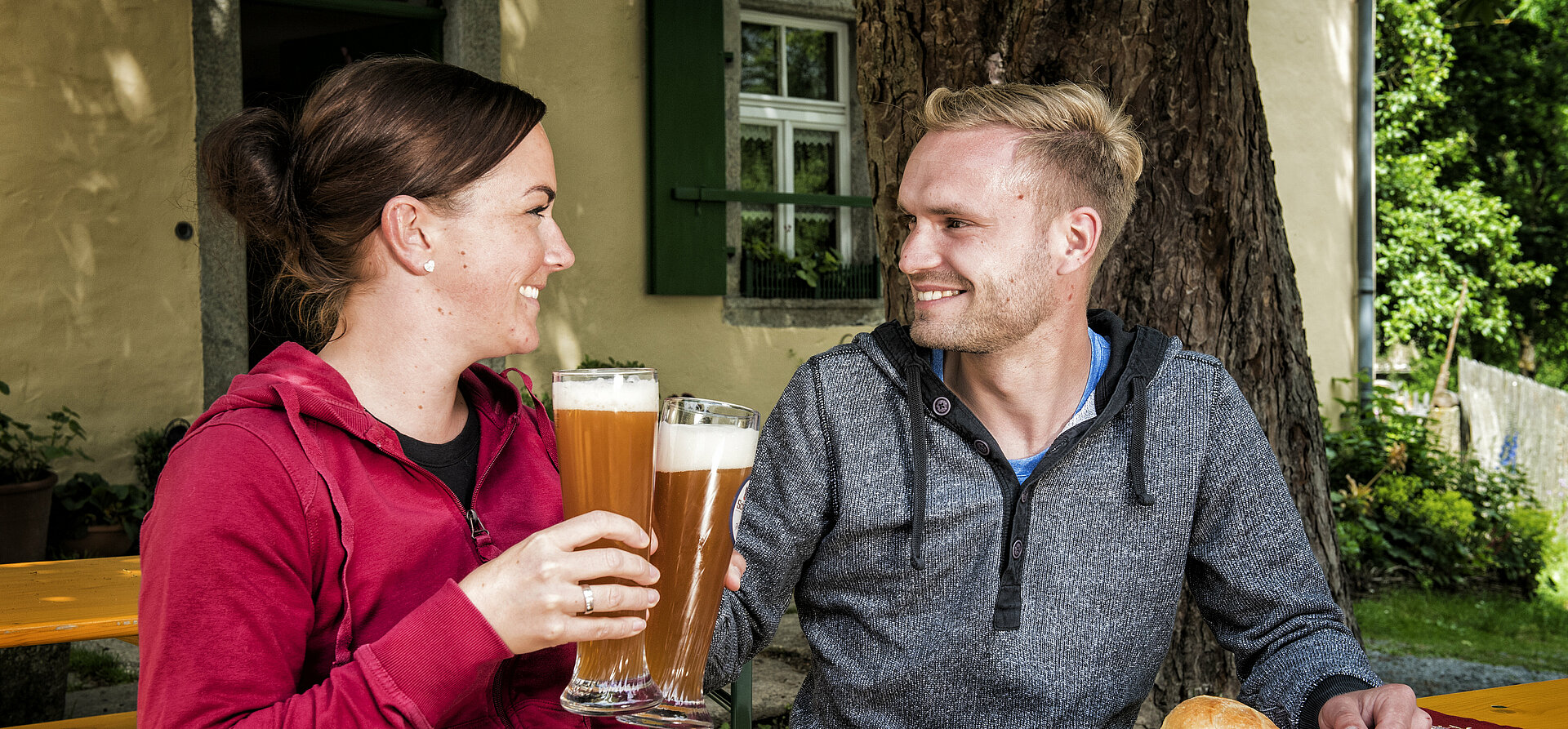 Ein Mann und eine Frau sitzen im Biergarten an einem gedeckten Tisch. Sie haben Biergläser in der Hand uns stoßen gerade an. 