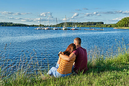 Ein Paar sitzt Arm in Arm am Seeufer. Auf dem See liegen Segelboote, im Hintergrund sind einige Schäfchenwolken am blauen Himmel zu sehen