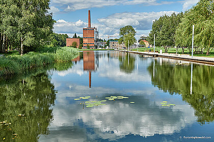 Wasserfläche mit Spiegelungen, im Hintergrund ein Industriedenkmal. Rechts und Links gesäumt von Bäumen 