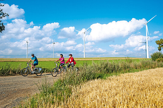 3 Radfahrer auf Radweg, im Vordergrund ein Kornfeld, im Hintergrund blauer Himmel und drei Windräder
