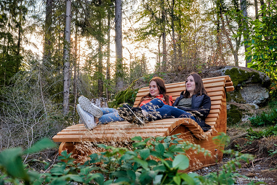 Zwei Frauen sitzen auf einer geschwungenen Holzliege im Wald.